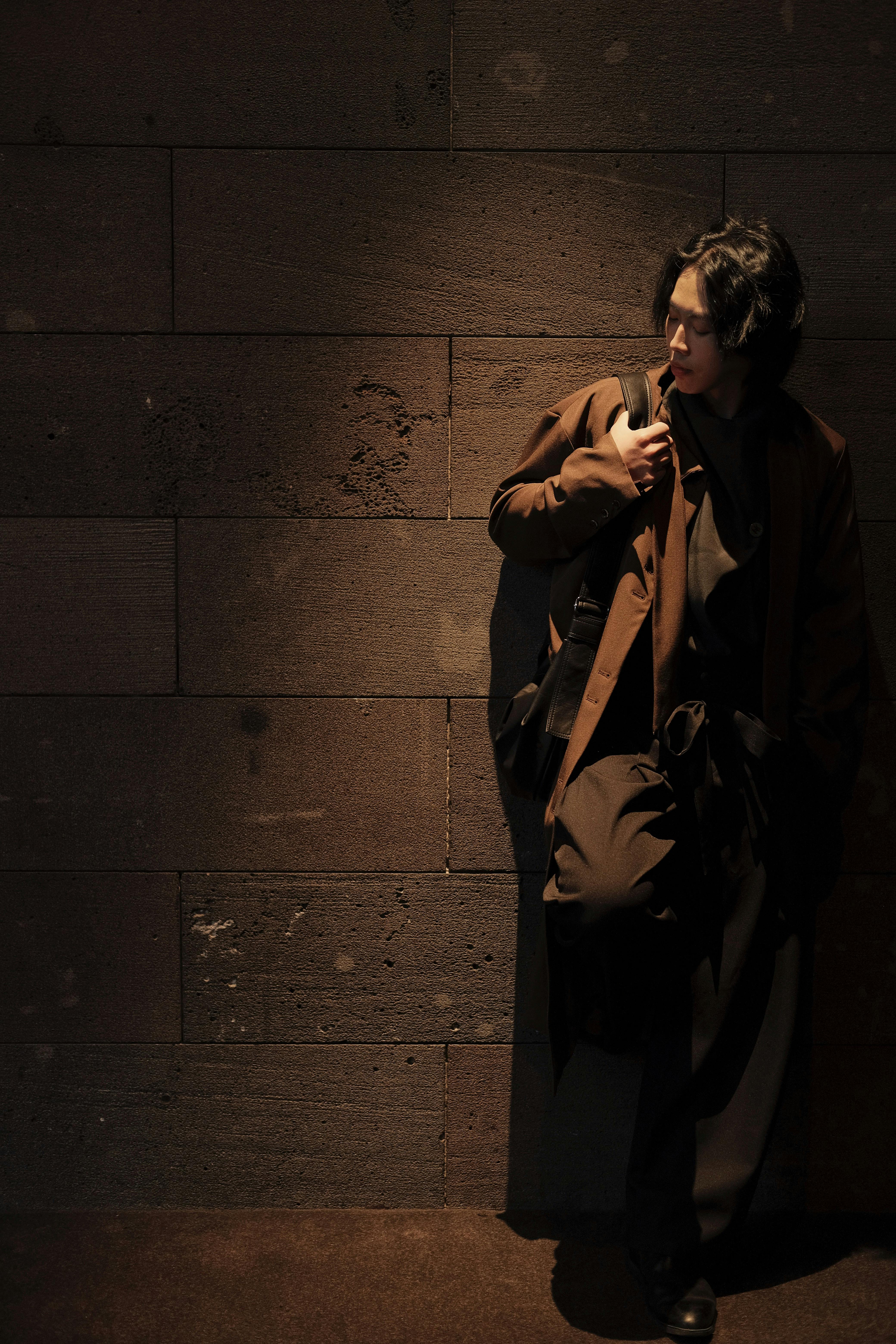 A fashionable person in a dark outfit leans against a textured wall, creating a moody urban scene.