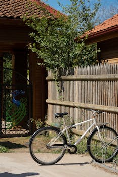 A bicycle rests against a rustic wooden fence beside a gate adorned with a peacock design on a sunny day.
