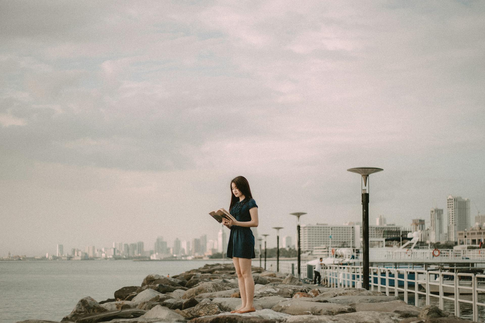 A woman stands reading on the rocky shore of Manila Bay with a city skyline in the background.