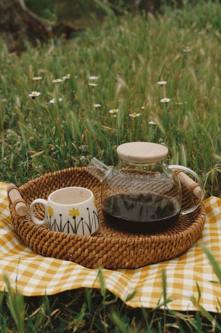 A cozy outdoor setup with a wicker tray holding a coffee pot and floral mug on a yellow checkered blanket in a grassy field.