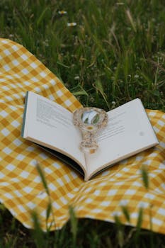An open book with a magnifying glass on a yellow gingham blanket in a grassy field.