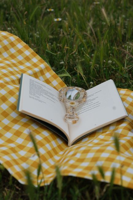 An open book with a magnifying glass on a yellow gingham blanket in a grassy field.