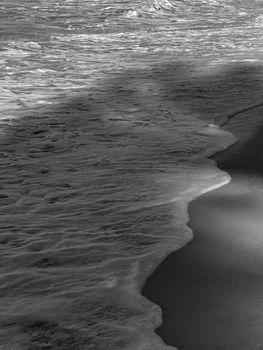 A dramatic black and white close-up of waves rolling onto a sandy beach.