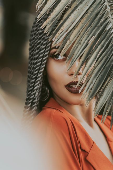A stylish woman in bold makeup poses behind palm leaves for a fashion shoot.