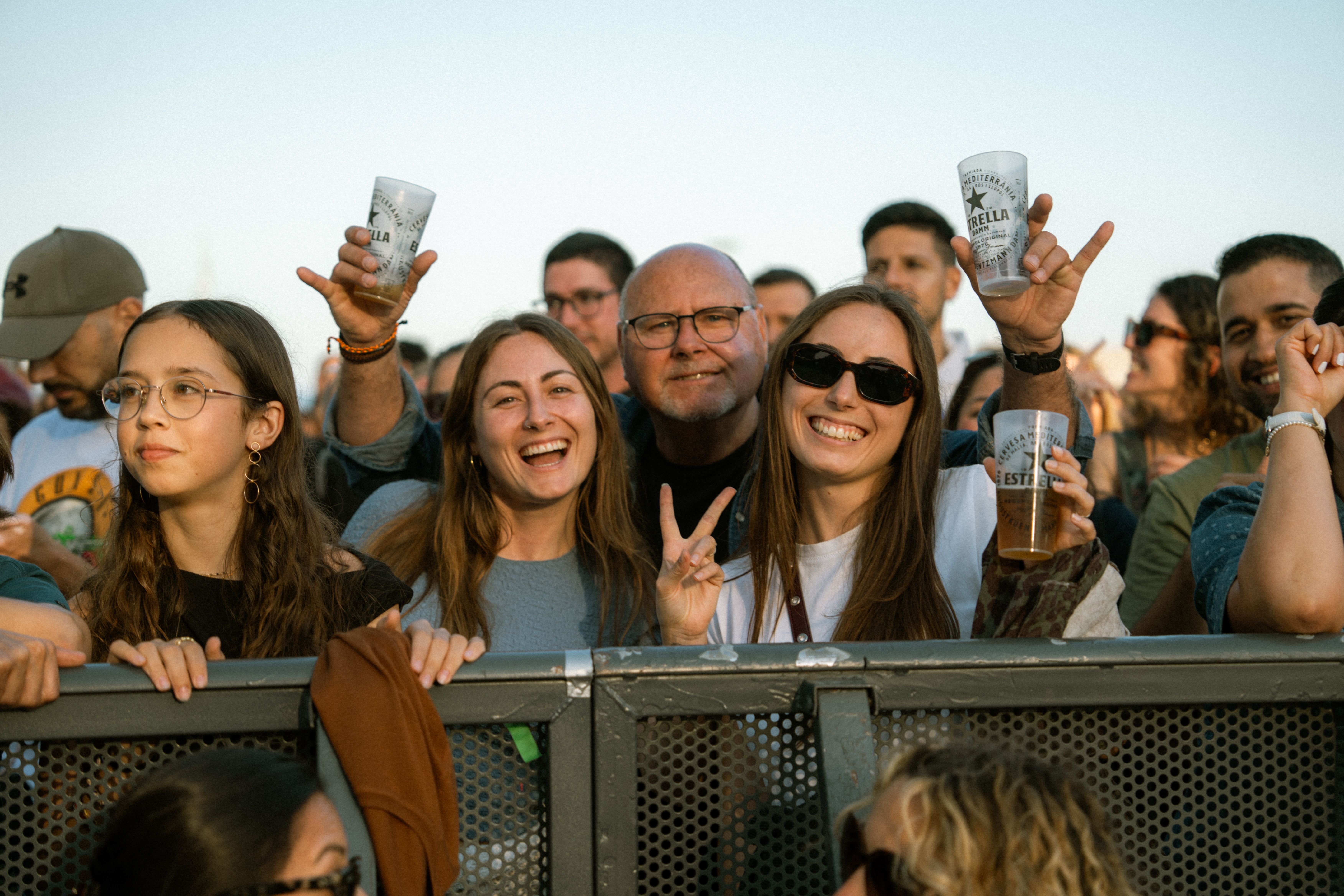 Joyful festival-goers enjoying live music, raising drinks, and showing peace signs at an outdoor event.