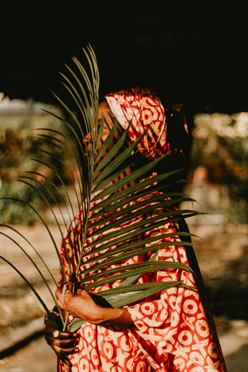 Free A woman in a red patterned dress holds a palm leaf outdoors, capturing a moment of cultural elegance. Stock Photo