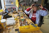 From above of cheerful female worker in headband magnifier glasses sitting at table with assorted tools and microchips while looking down