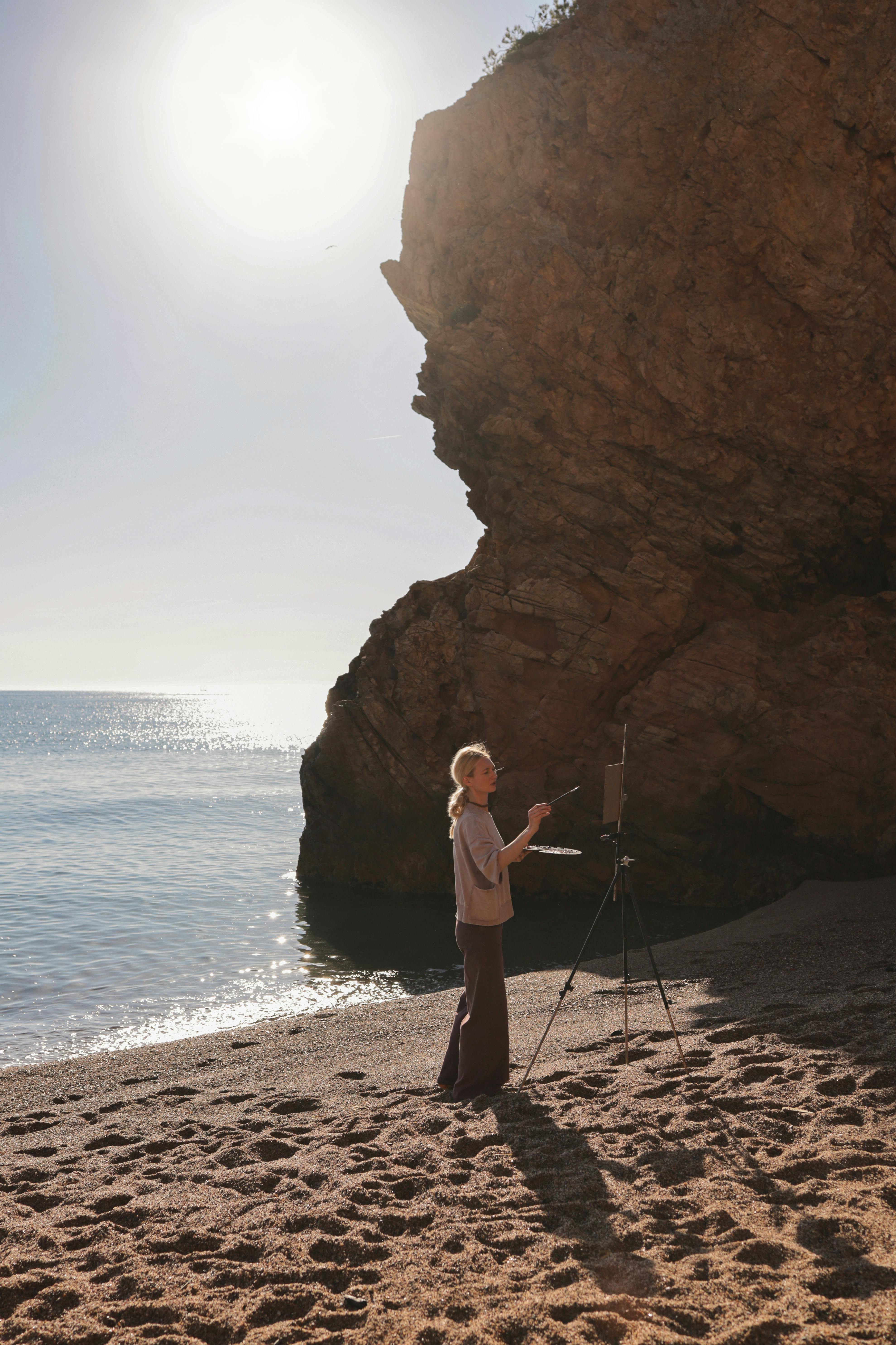 A person paints a seascape on a sunny beach with dramatic cliffs.