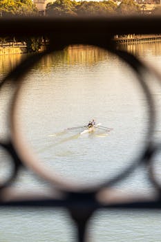 Two people rowing on the Guadalquivir River in Seville, framed artistically by a railing.