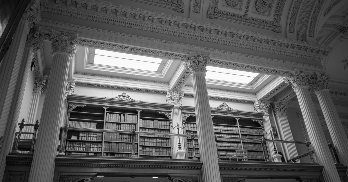 A black and white photo of a grand library interior with ornate columns and filled bookshelves.