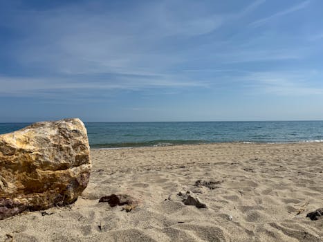 Serene beach view with a large rock, blue sky, and calm ocean waves.