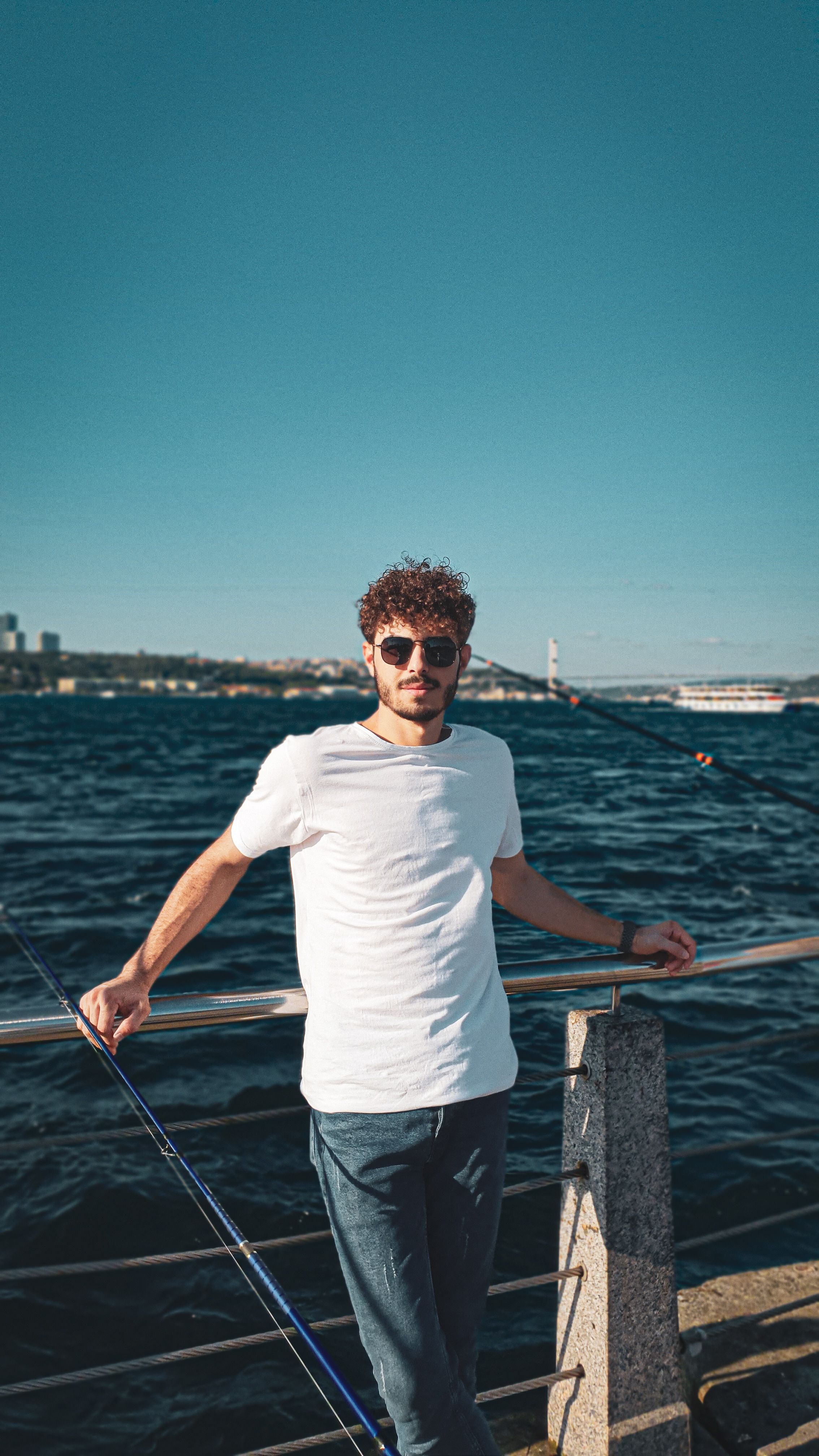 A young man stands by a waterfront railing enjoying a sunny day with a city in the background.