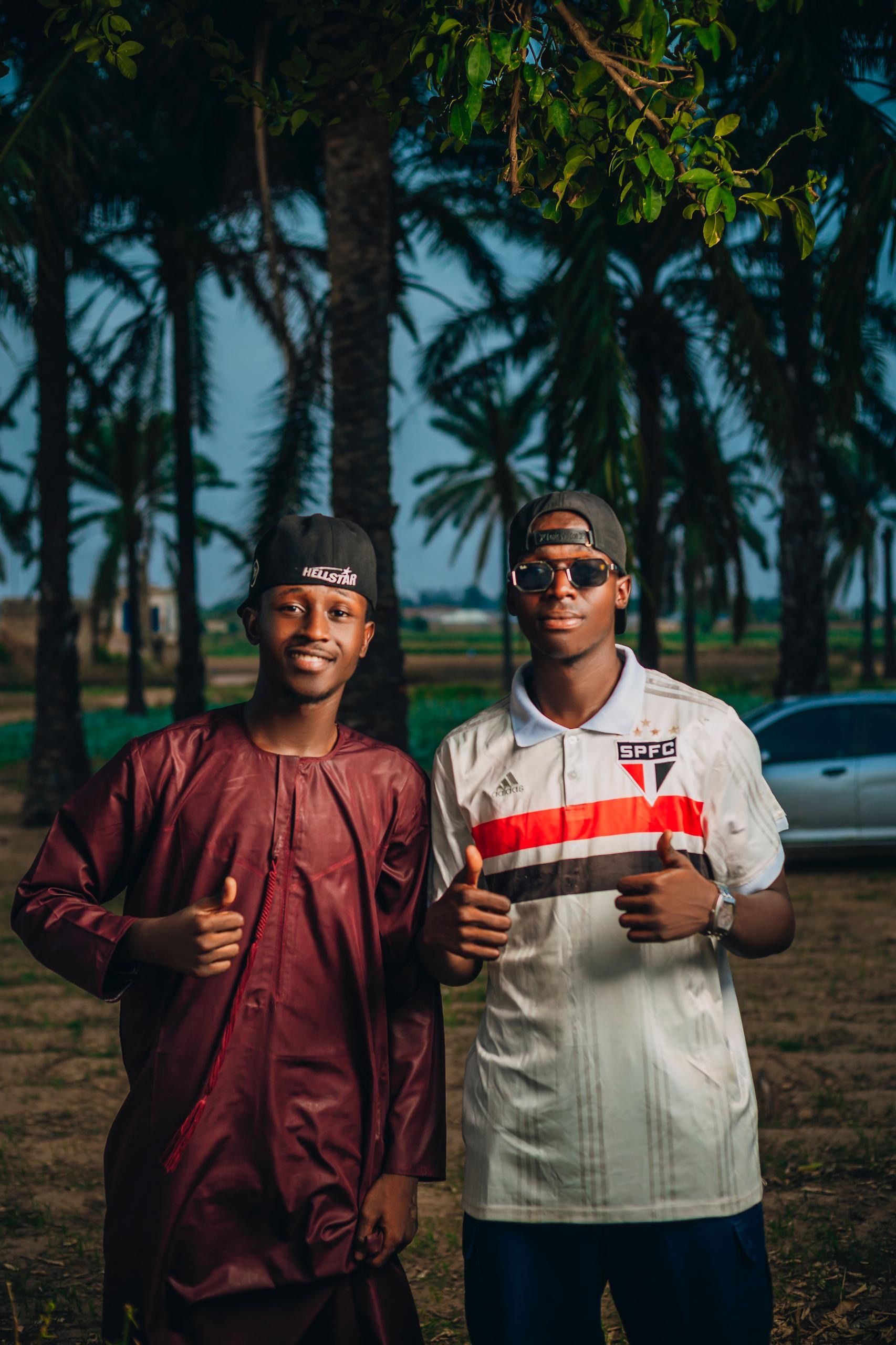 Two young men giving a thumbs up, standing outdoors among palm trees.