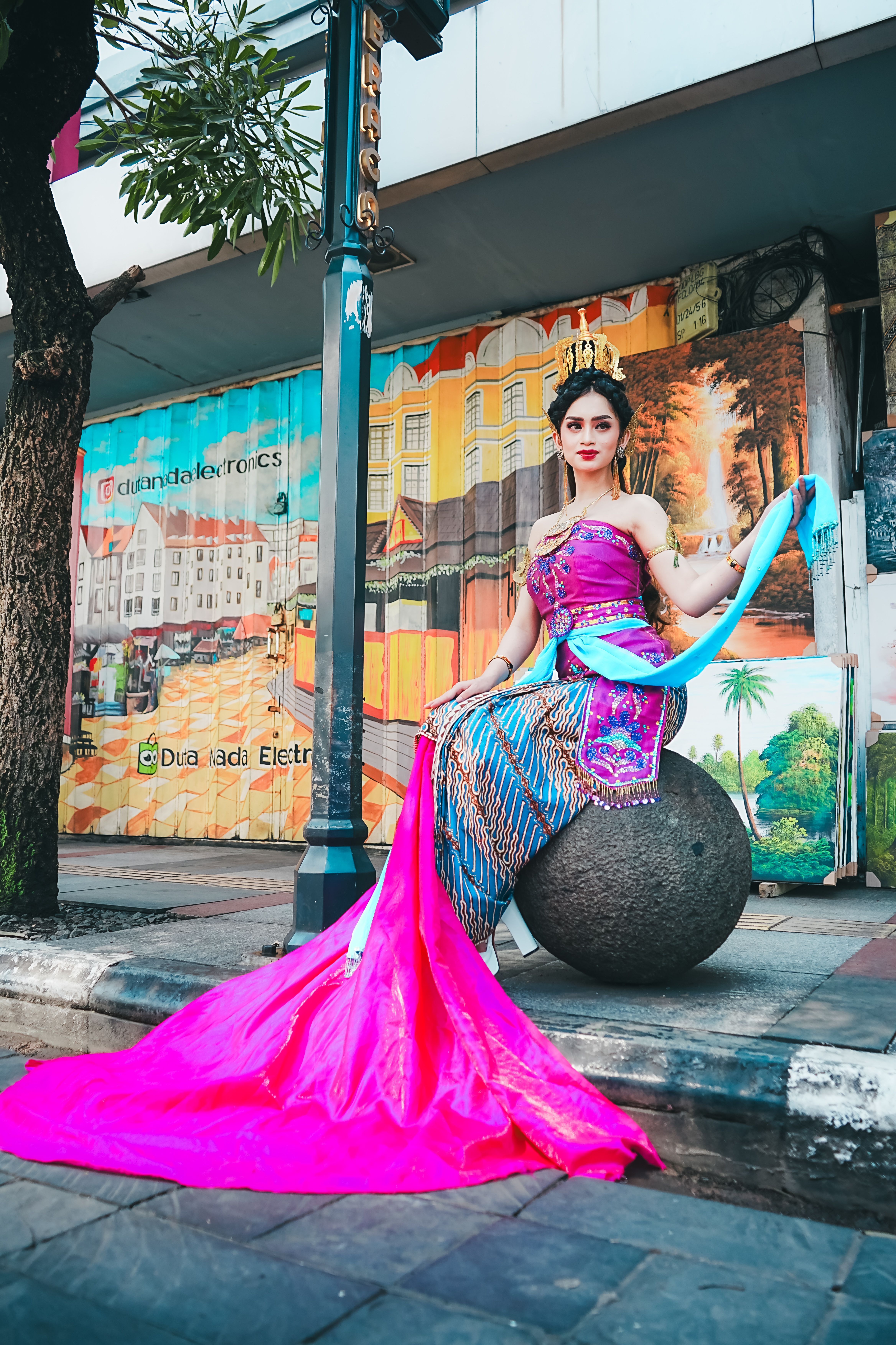 A woman in vibrant traditional dress sits gracefully on a sphere in an urban setting, surrounded by colorful murals.