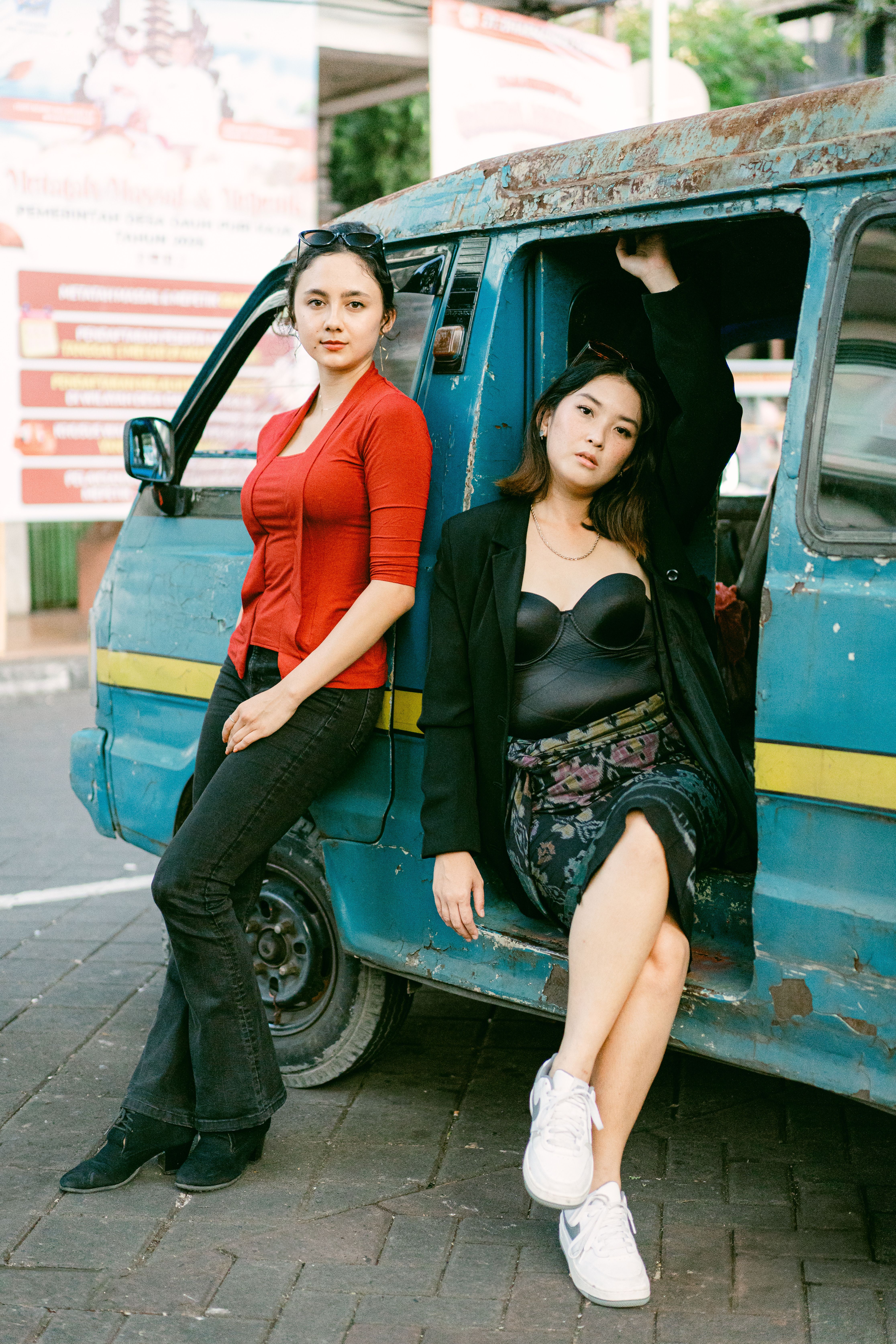 Two stylish young women posing confidently by a vintage blue van in an urban outdoor setting.