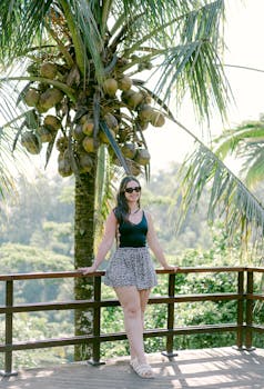 Woman standing by railing under a palm tree, enjoying a sunny outdoor setting.