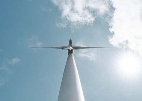 A striking view of a wind turbine from below, set against a vibrant sky with sun and clouds.