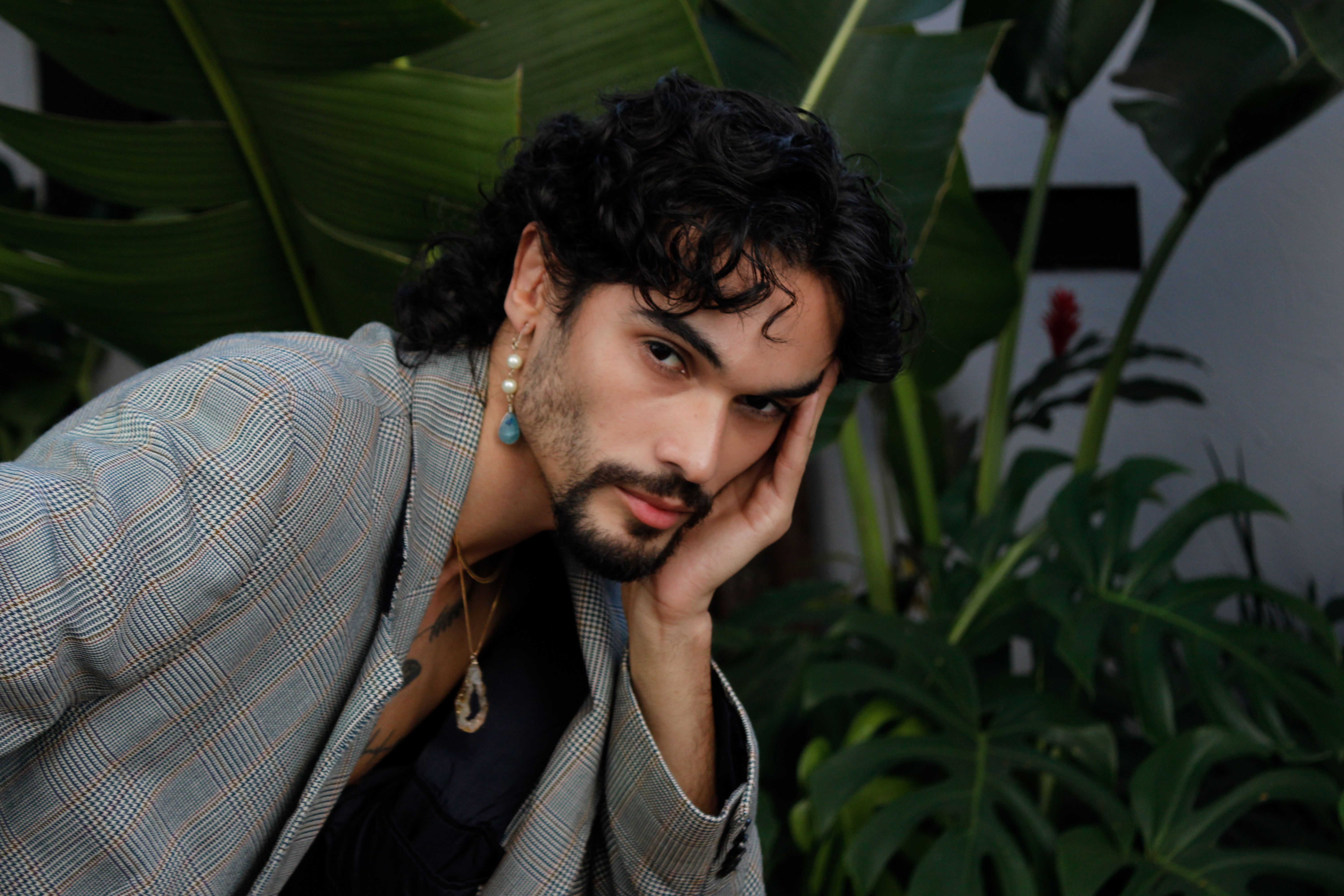 Man with curly hair in fashionable outfit, surrounded by lush tropical plants, conveying a relaxed mood.