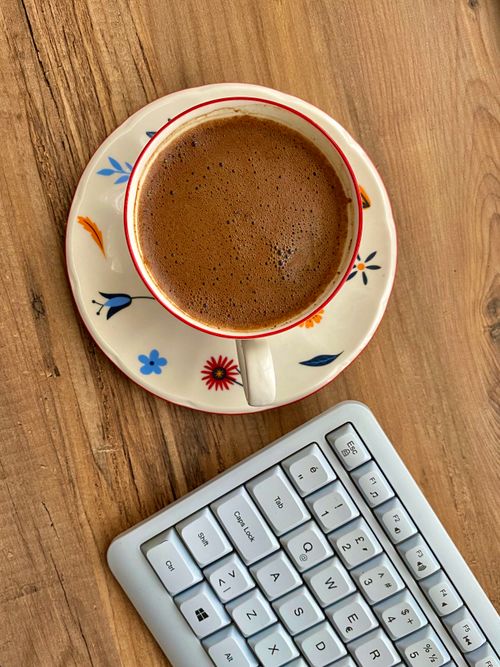 Free Top view of a coffee cup on a floral saucer next to a keyboard on a wooden desk. Stock Photo