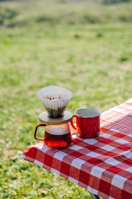 Camping coffee maker setup with a kettle, pour-over brewer, enamel mugs, and a sealed coffee container on a picnic table