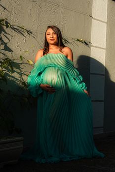 Pregnant woman in flowing green dress poses outdoors with soft sunlight. - Fotografer Sidoarjo