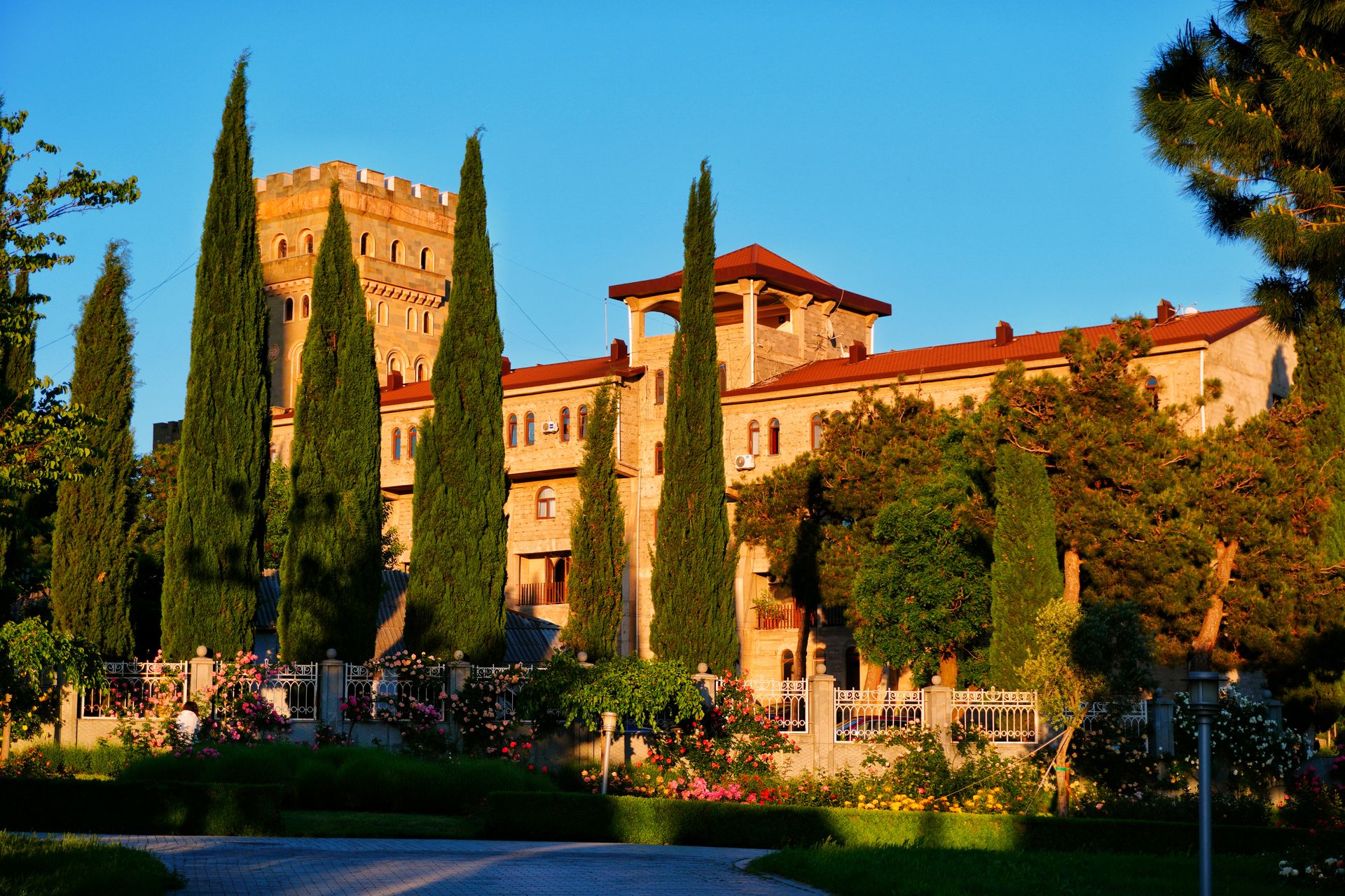 A grand Mediterranean-style castle surrounded by lush gardens and tall cypress trees under a bright sky.