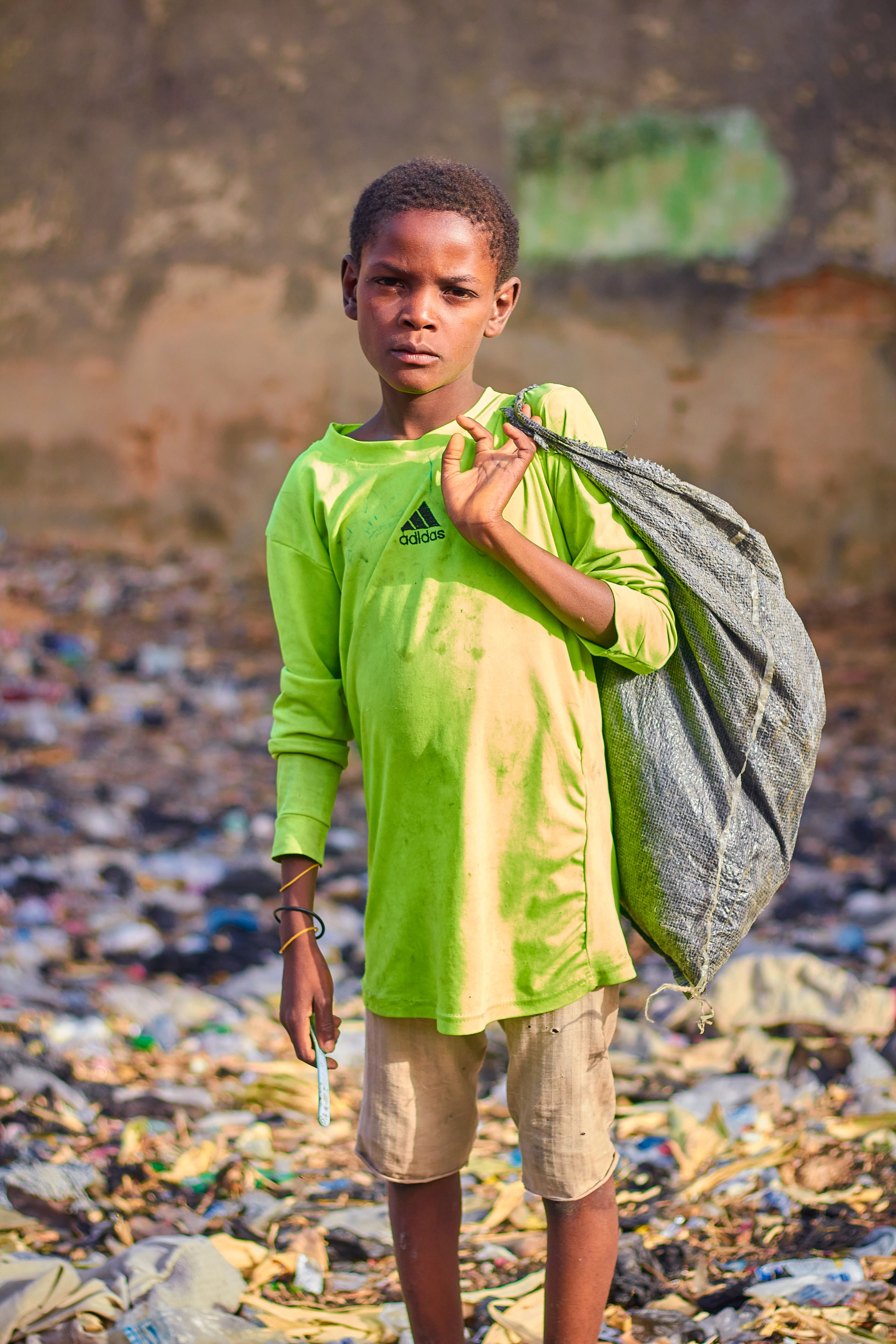 Child in lime green shirt holding a bag standing in a debris-filled area.