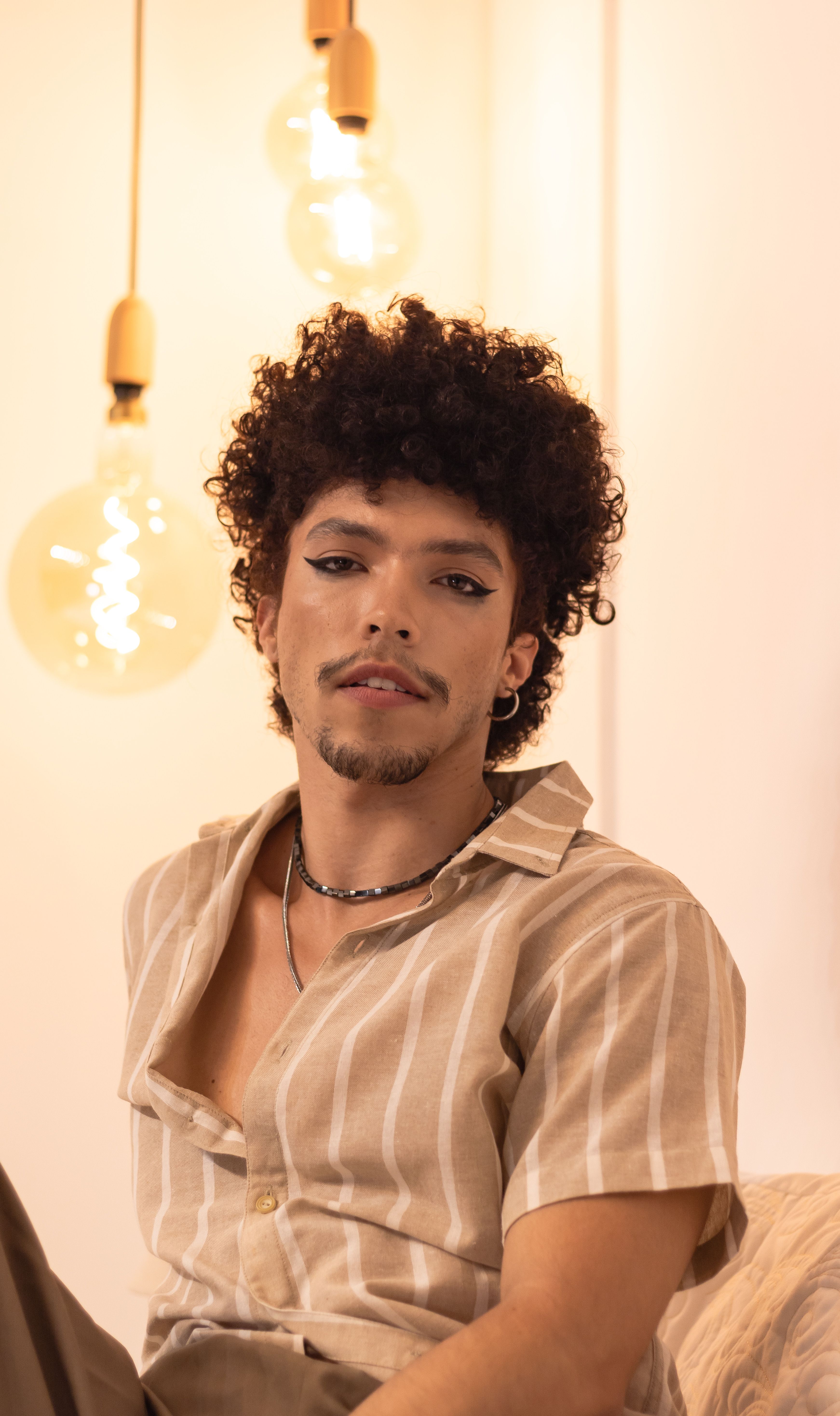Young man with curly hair and makeup in a casual indoor setting with warm lighting.