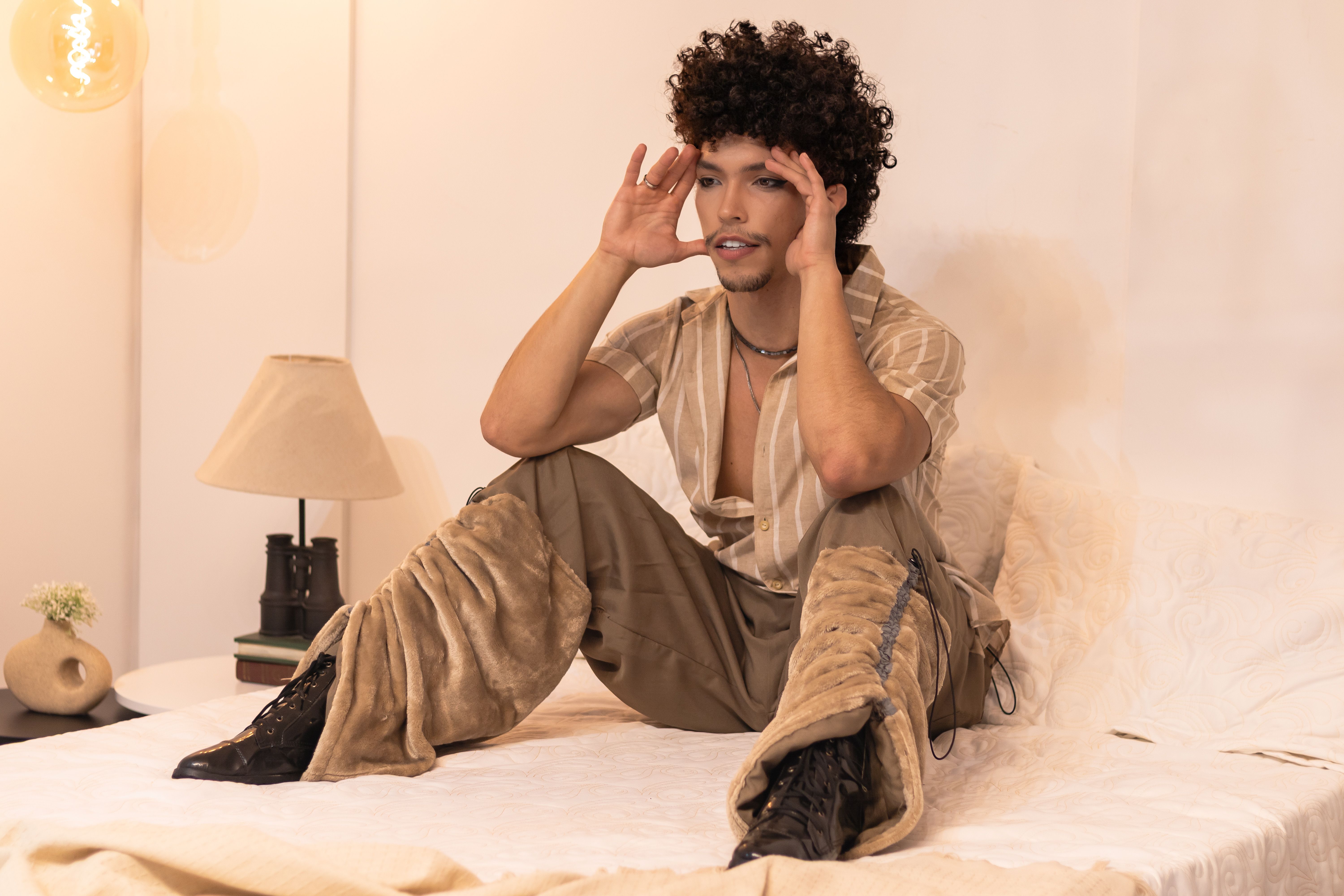 Young adult seated on bed with modern fashion attire, warm lighting.