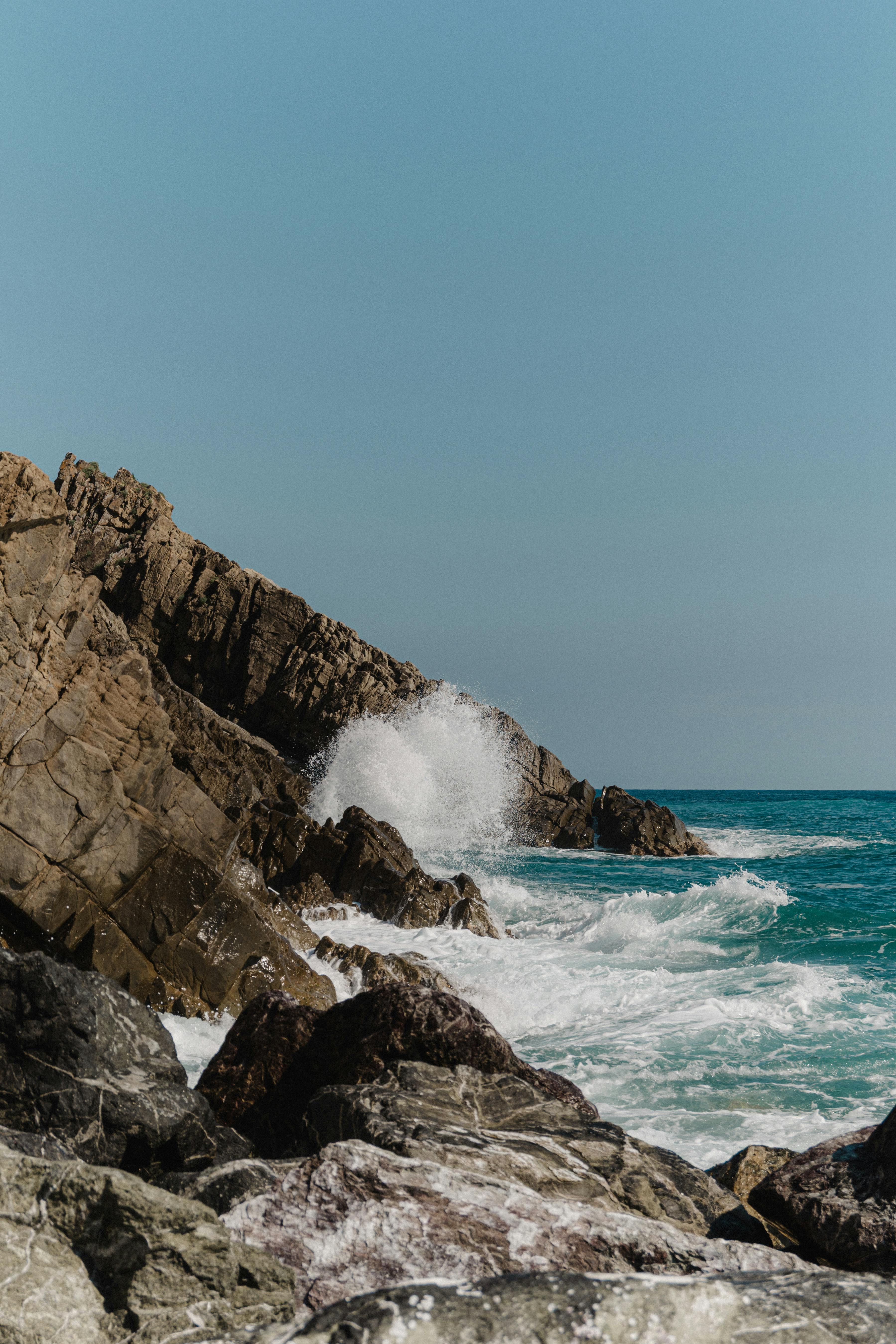 Waves crash against a rugged rocky coastline under a clear sky, showcasing natural beauty.