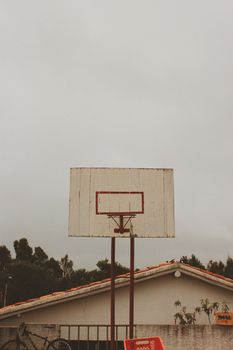 Old basketball hoop with vintage backboard stands outdoors on a cloudy day, bordered by foliage.