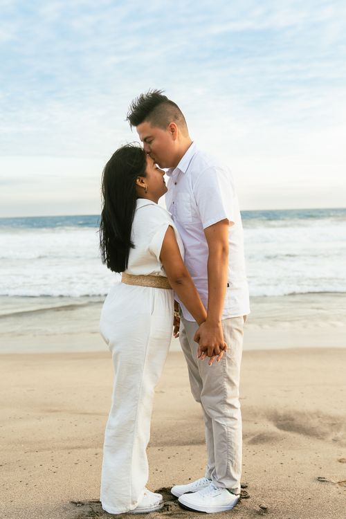Free A couple in white attire shares a romantic kiss on a serene beach at sunset. Stock Photo