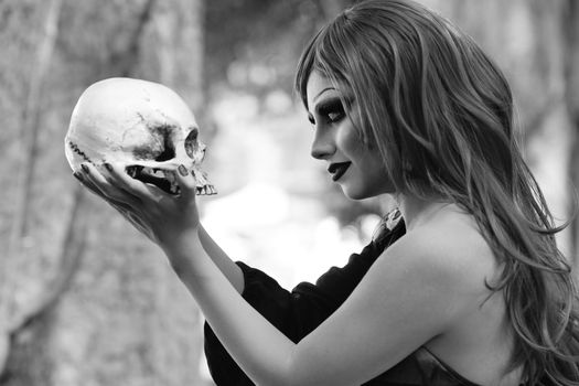 Dramatic black and white portrait of a woman with artistic makeup holding a skull in an outdoor setting.