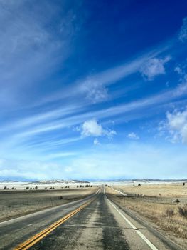 A long highway stretches through vast open plains beneath a vivid blue sky with wispy clouds.