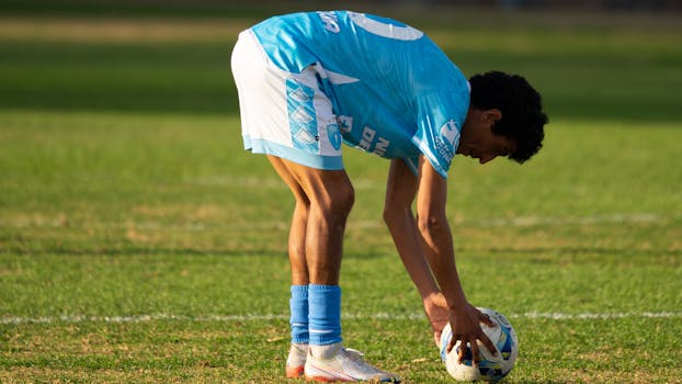 A soccer player in a blue uniform prepares to kick a ball on a grass field during the day.