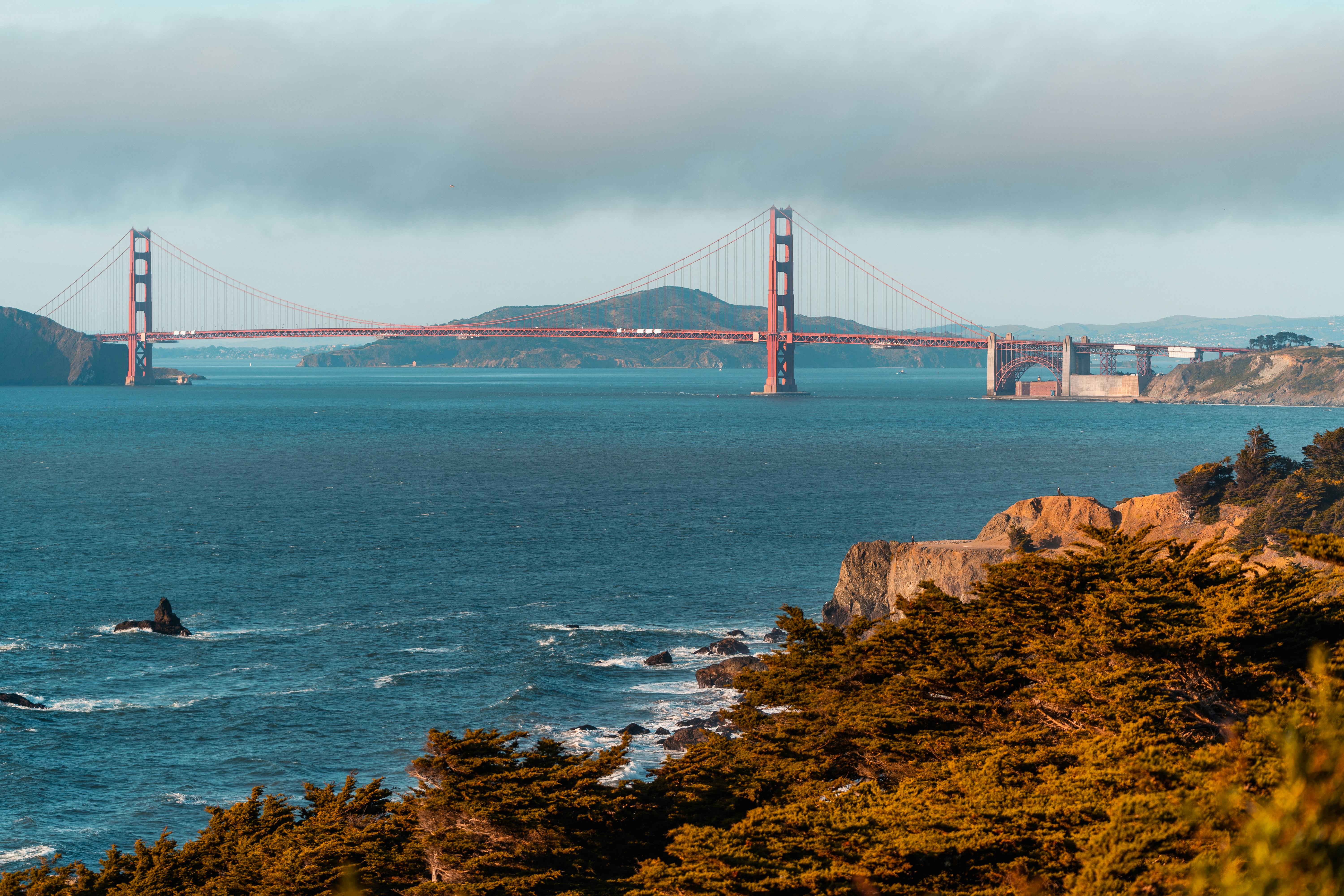 Scenic view of the Golden Gate Bridge with ocean and lush trees under a cloudy sunset sky.