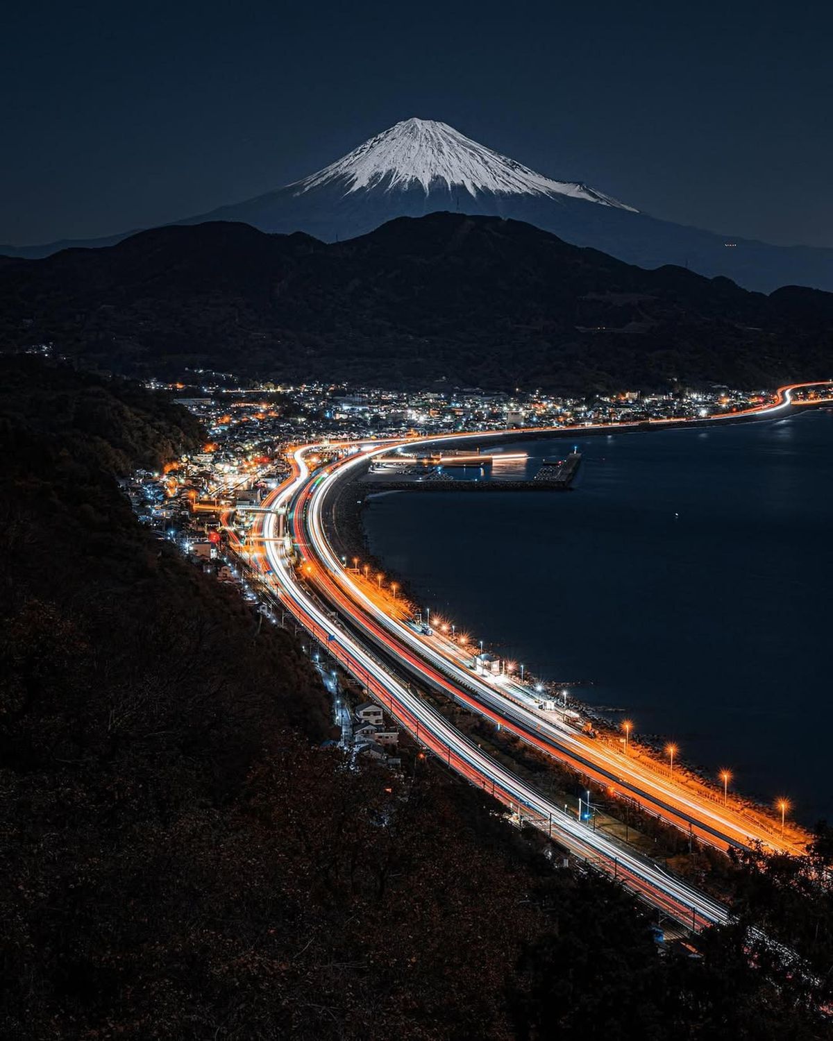 Mount Fuji at night above Shizuoka city lights and Suruga Bay coastline — the south face of the volcano and the Izu Peninsula coast lit at night