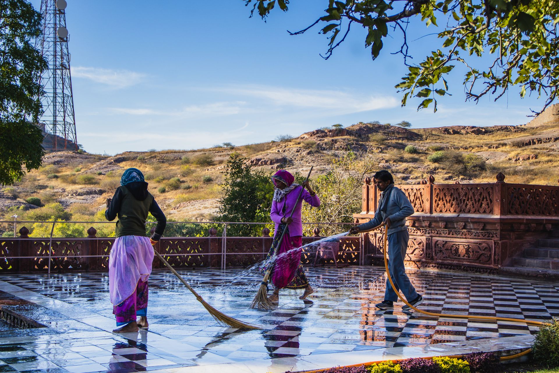 Three individuals cleaning an outdoor tiled courtyard in a scenic Indian landscape under a clear blue sky.