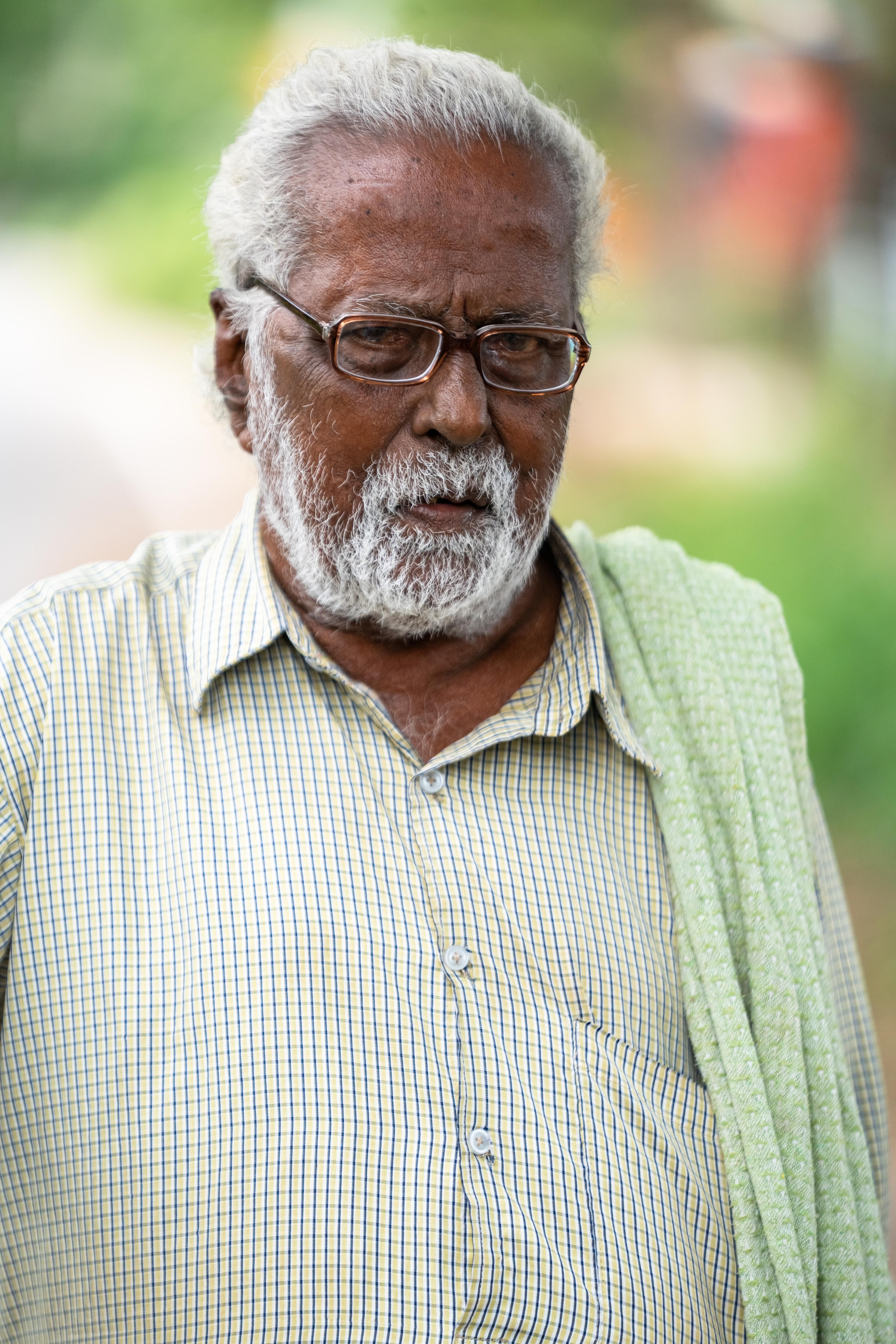Portrait of an elderly man outdoors, wearing glasses and a casual shirt, with a thoughtful expression.