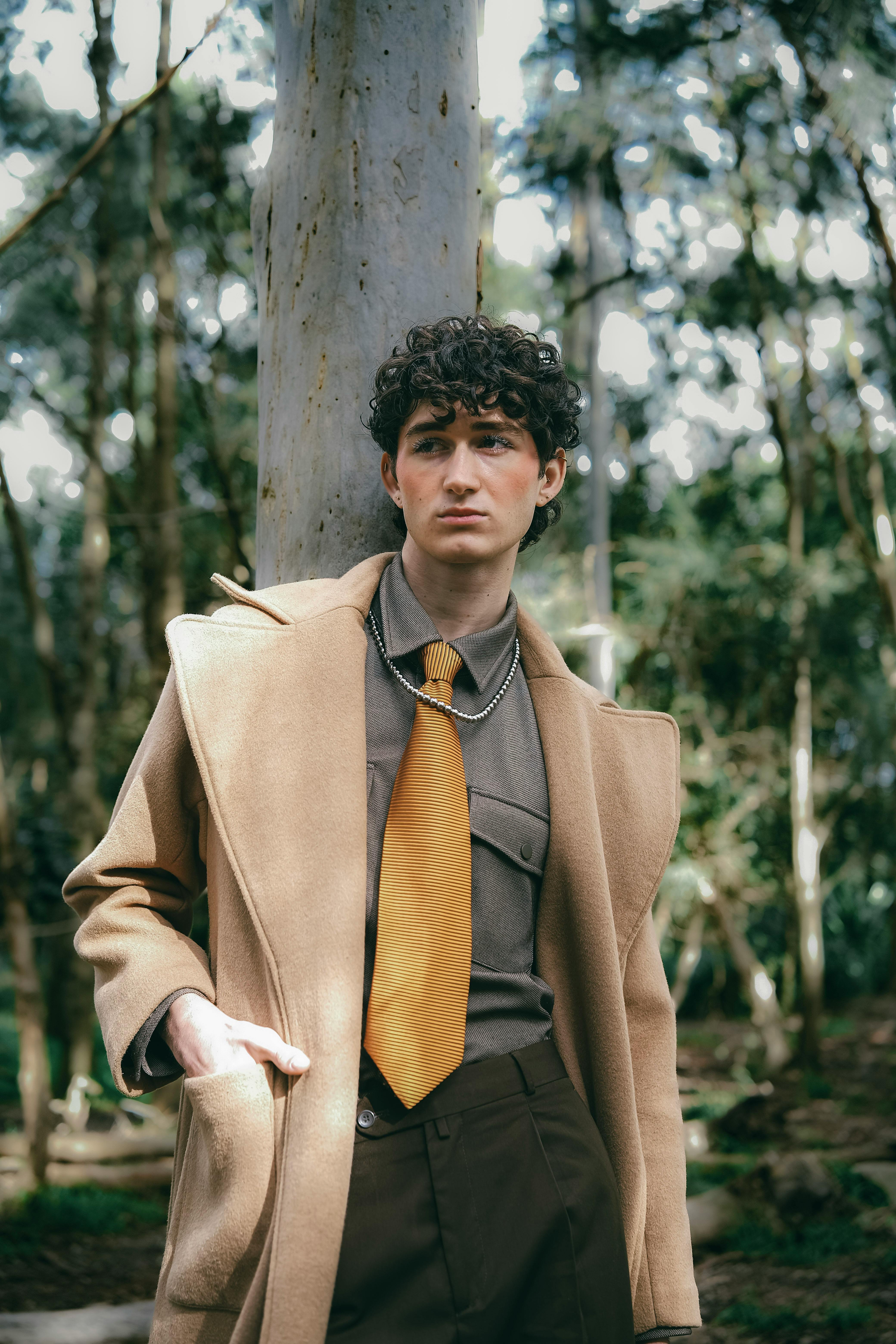 Young man in a fashionable coat stands confidently in a lush forest, exuding elegance.