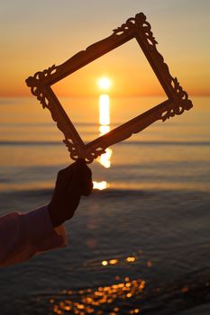 Artistic sunset view framed by hand, highlighting calm ocean waves and glowing sun.