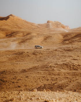 A lone SUV navigates a vast desert, raising dust under a clear blue sky.