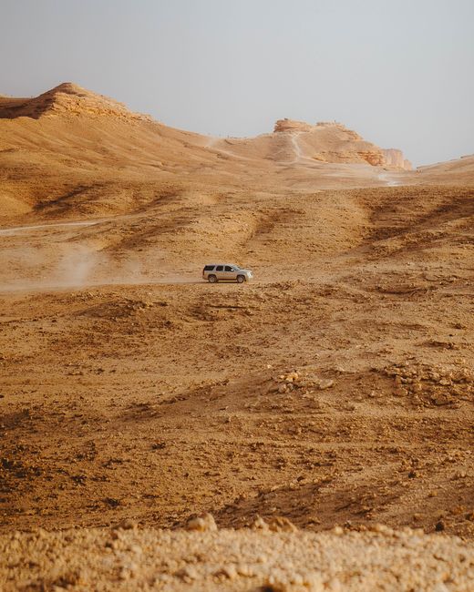 A lone SUV navigates a vast desert, raising dust under a clear blue sky.