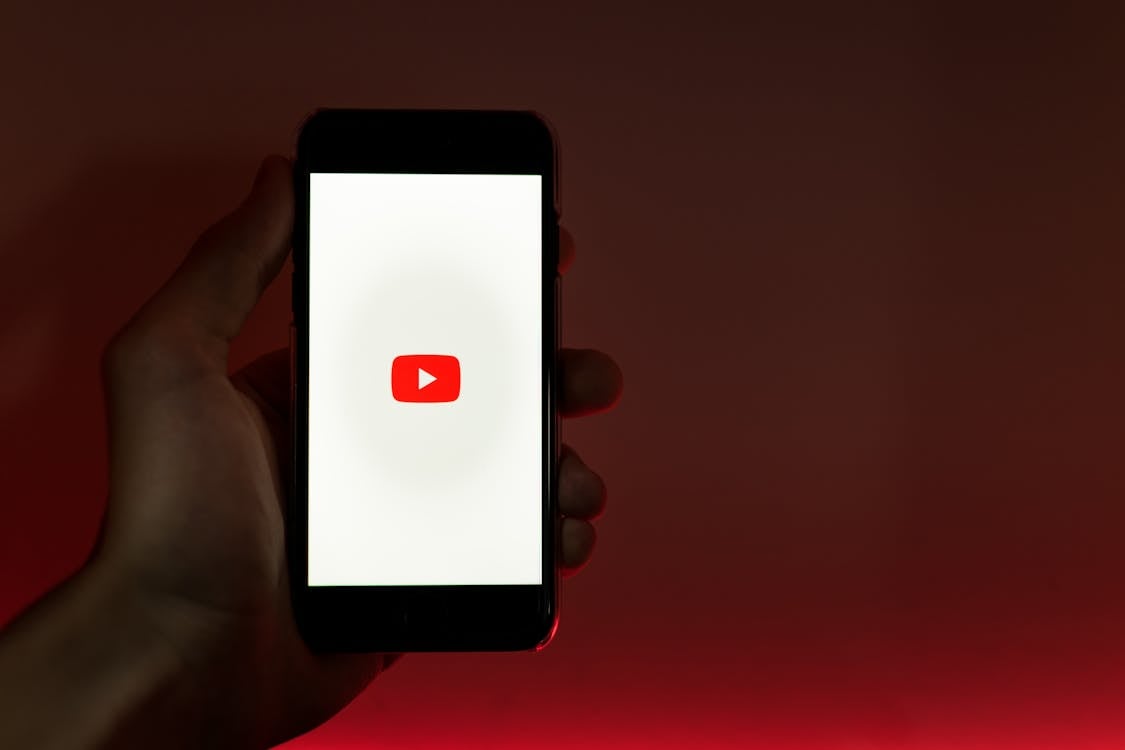 An image of a phone with the YouTube logo on the screen being held by a left hand on a red background. 