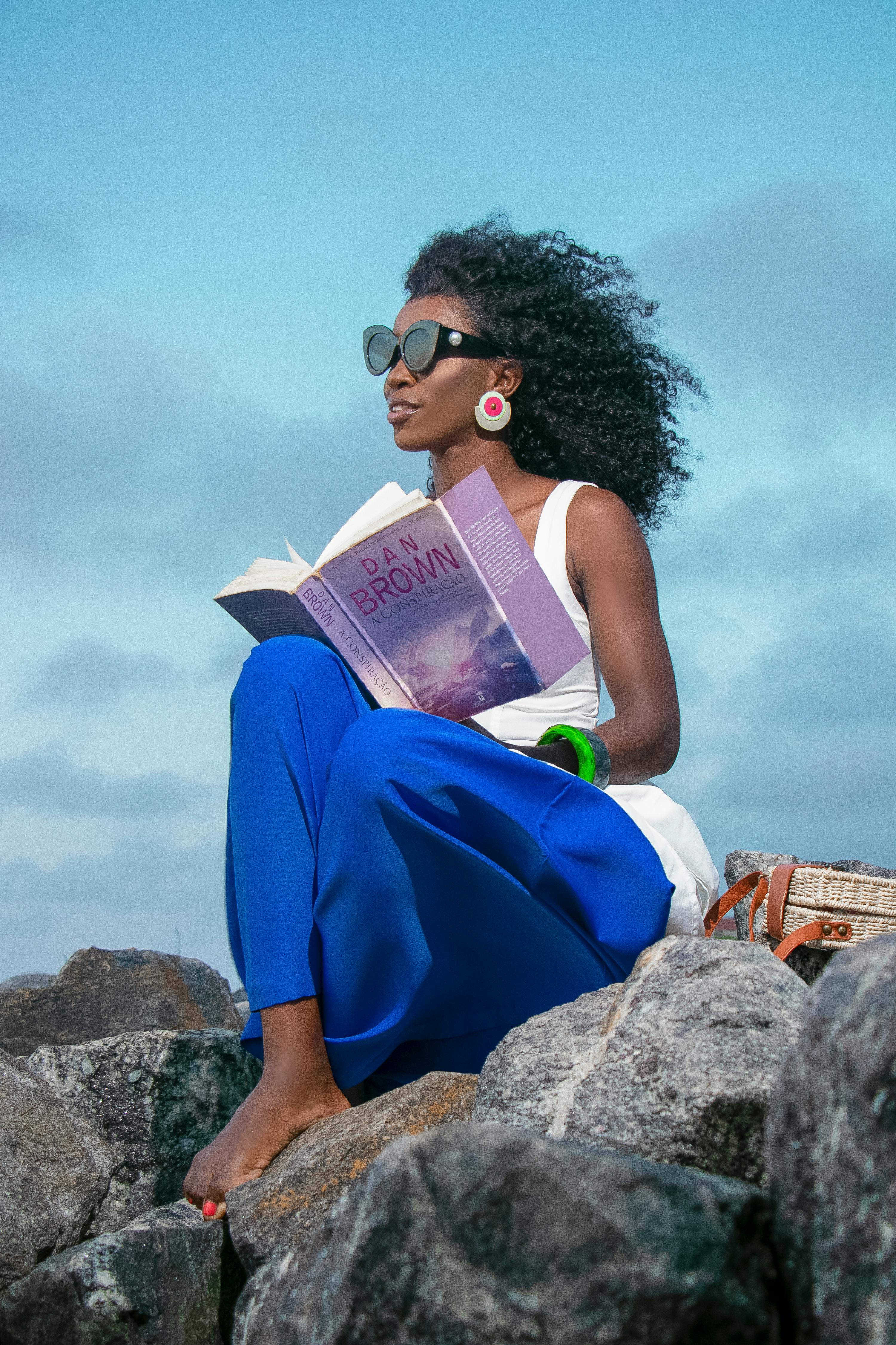 Elegant woman reading a novel while sitting on rocks showcasing fashion and leisure.