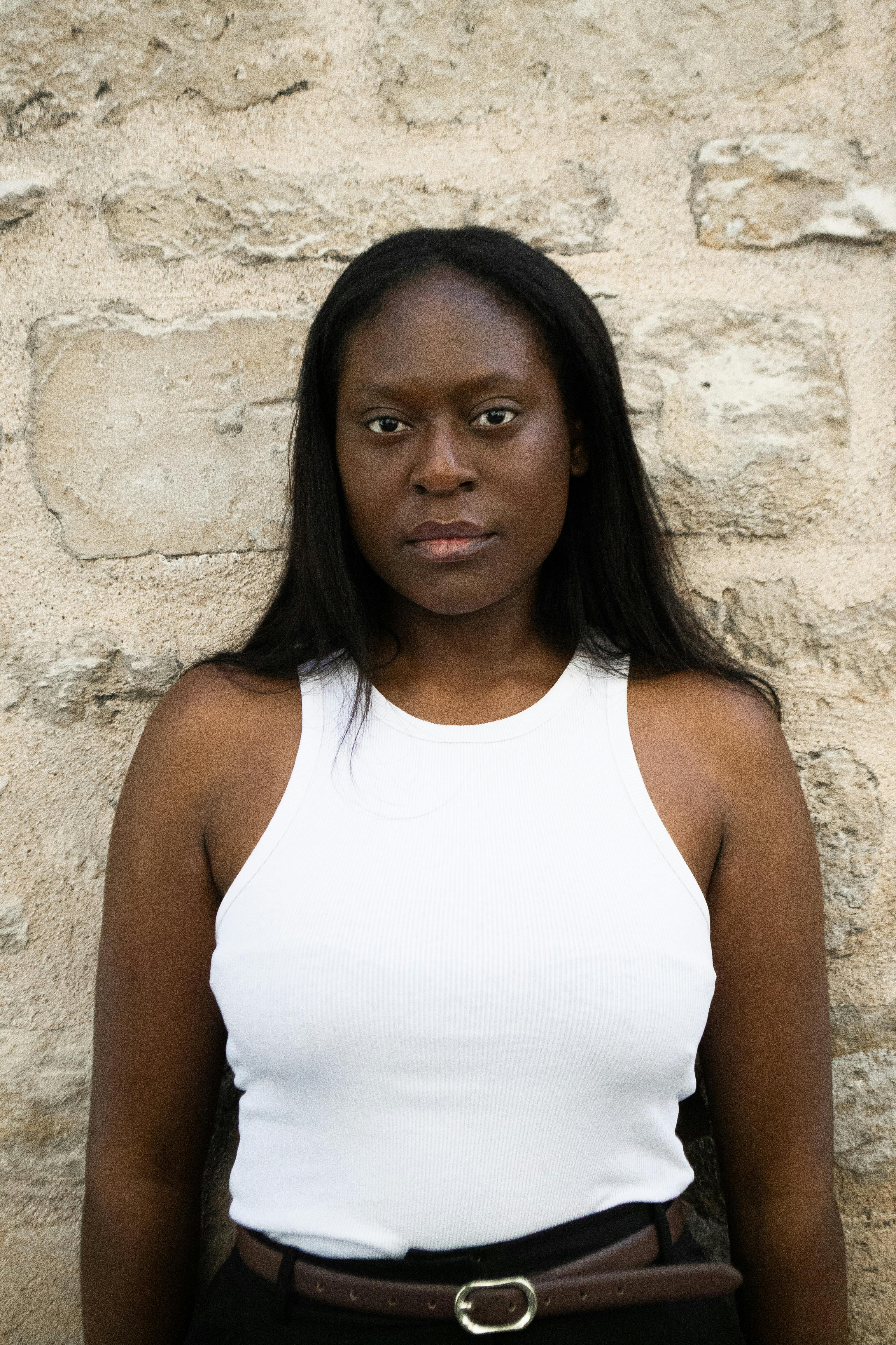 Confident woman in a white tank top stands against a rustic stone wall.