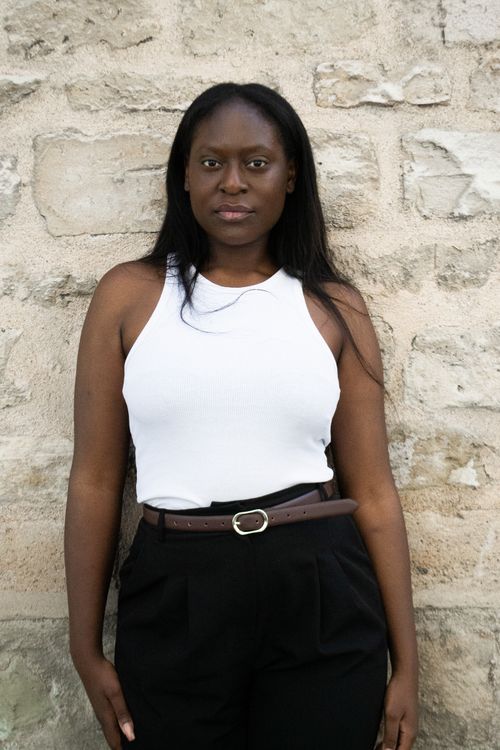 Free Portrait of a confident woman standing against a rustic stone wall, wearing white top and black pants. Stock Photo