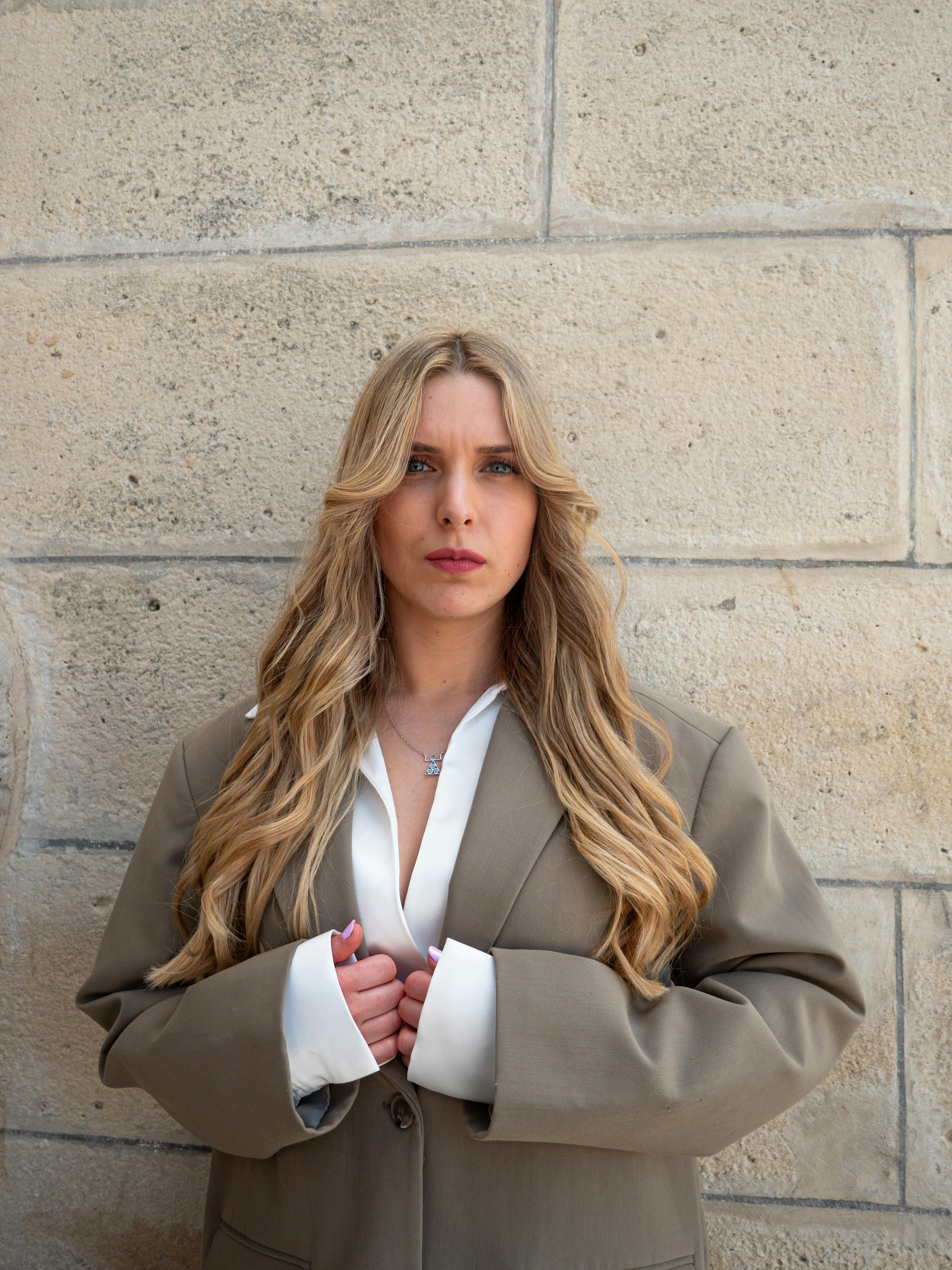 Blonde woman in a beige jacket stands confidently against a textured stone wall, showcasing elegance and style.