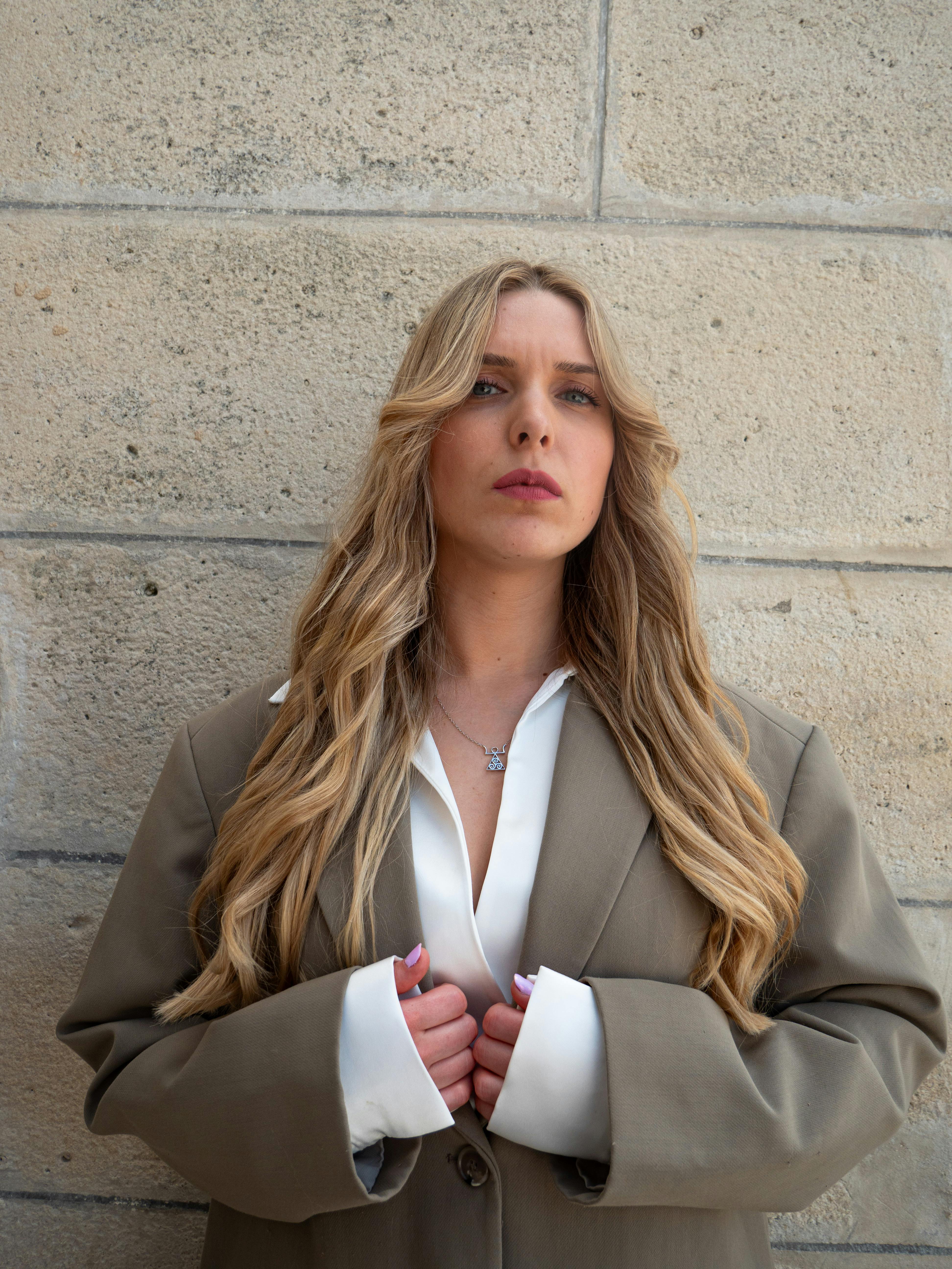 Young woman with long hair poses in a blazer against a textured stone wall, exuding modern elegance.