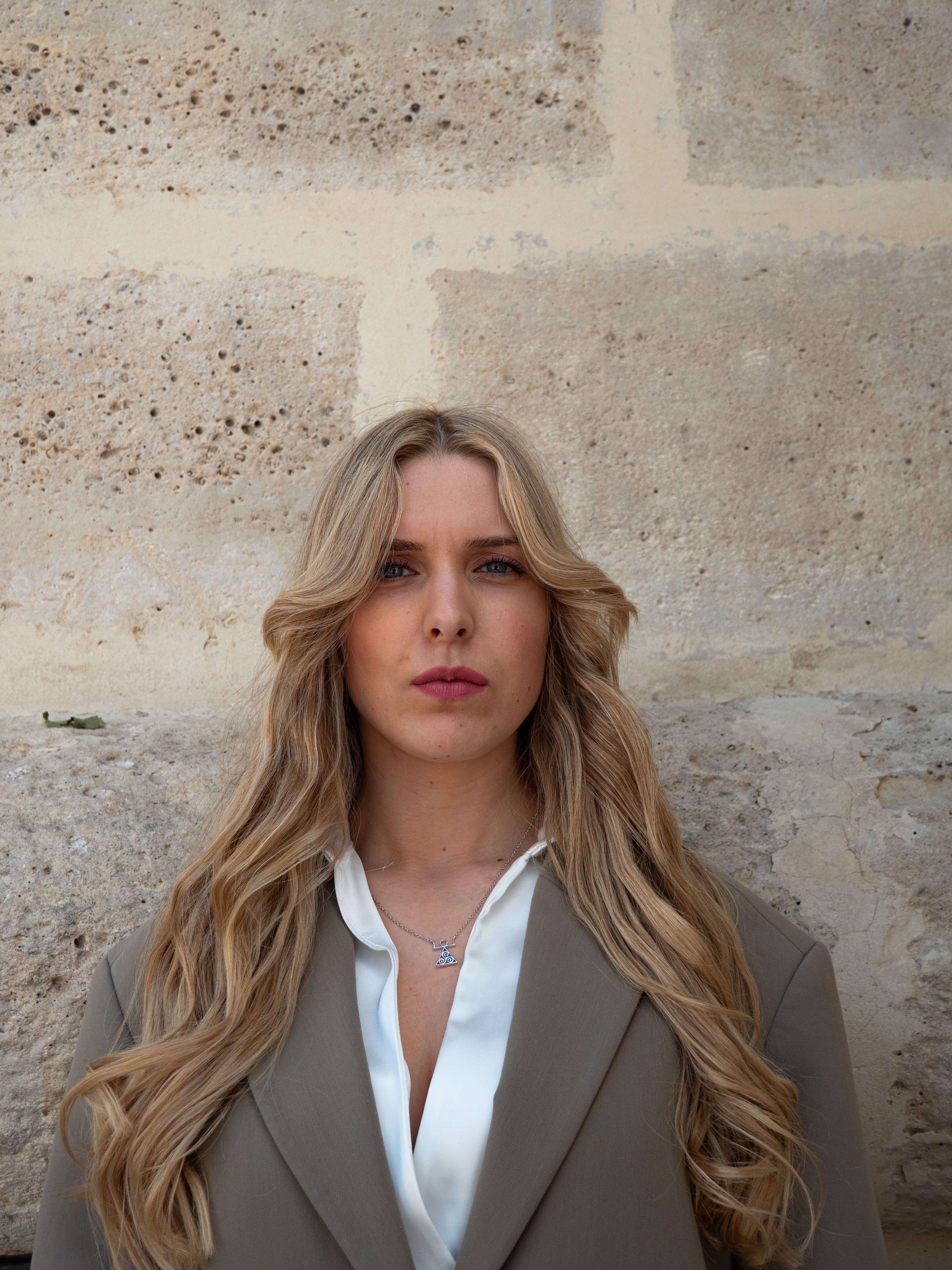 Portrait of a woman with long blonde hair in a blazer against a textured stone wall.
